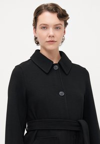 Giovane con capelli mossi e corti che indossa orecchini a cerchio e un cappotto nero con cintura e due bottoni, su uno sfondo chiaro e semplice.