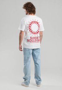 White cotton t-shirt features a red sun graphic and text "SUNSET BOULEVARD" at the back; paired with light blue denim jeans.
