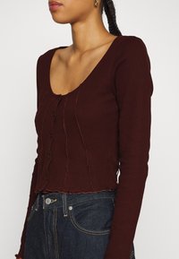 Top burdeos de manga larga con acanalado y escote redondo, con una fila de botones oscuros y un dobladillo ondulado; combinado con jeans de mezclilla oscuros.