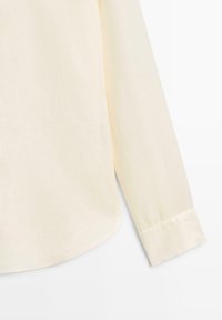 Crèmewit overhemd met lange mouwen, manchet en zoom met zichtbare stiksels, afgebeeld tegen een witte achtergrond.