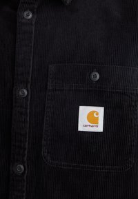 Chemise en velours côtelé noire avec une poche avant arborant un écusson logo Carhartt orange et blanc ainsi que des boutons noirs sur le devant.