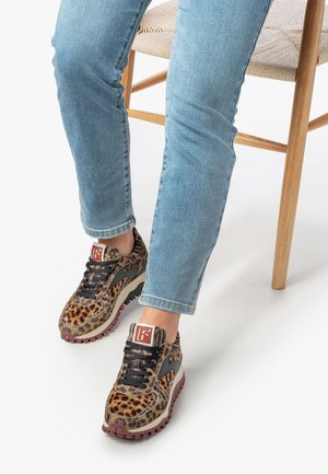Leopard-print sneakers with a fur texture, black laces, and a rubber sole. Paired with light blue denim jeans, seated on a chair.
