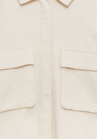 Beżowa bawełniana koszula z kołnierzykiem, dwiema przednimi kieszeniami i fakturowanym materiałem. Projekt jest prosty, z widocznymi szwami.