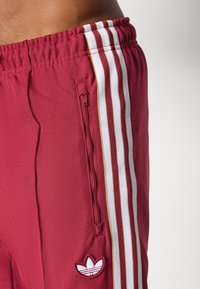 Sarkani sporta šorti ar baltām un zelta vertikālām svītrām, elastīgu vidukli, sānu rāvējslēdzēja kabatu un mazu baltu logotipu apakšējā kreisajā pusē.