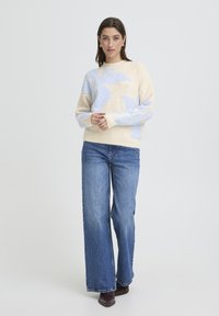 Beige neulepusero, jossa vaaleansininen lehtikuvio, yhdistetty leveälahkeisiin sinisiin farkkuihin ja tummanruskeisiin kenkiin. Sileä pinta ja resorividat.