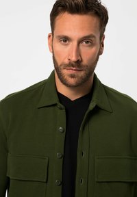 Groene gestructureerde jas met een kraag, twee voorzakken en knoopsluiting, gelaagd over een zwarte V-hals shirt.