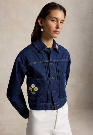 Žena v tmavomodrej rifľovej bunde s kvetinovou výšivkou na vrecku a golieri, v bielych nohaviciach, stojaca pred béžovým pozadím.