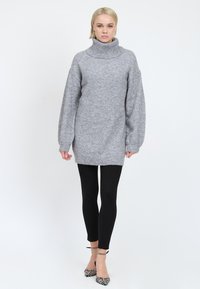 Grå strik turtleneck sweater med lange, voluminøse ærmer, ribbet kant, båret over sorte leggings og parret med mønstrede hæle.