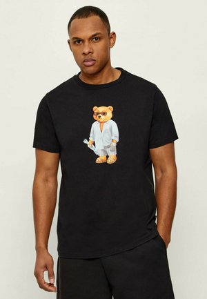 Uomo che indossa una maglietta nera con un orso con occhiali da sole, accappatoio bianco, pantaloncini a righe, sandali e tiene una bottiglia.