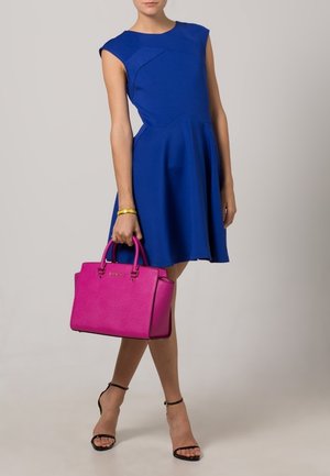 Kobieta ubrana w niebieską sukienkę bez rękawów, czarne szpilki z paskiem wokół kostki, trzymająca jasnoróżową torebkę, z złotą bransoletką na nadgarstku.