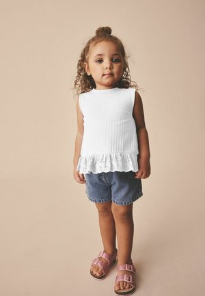 Kleinkind mit lockigem Haar in einem weißen ärmellosen Top mit Rüschen, Jeansshorts und pinken Sandalen steht vor einem beigen Hintergrund.