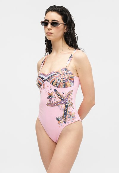 Femme portant un maillot de bain une pièce rose avec des motifs colorés d'étoile de mer en perles et de coquillages, ainsi que de petites lunettes de soleil, debout devant un fond uni.