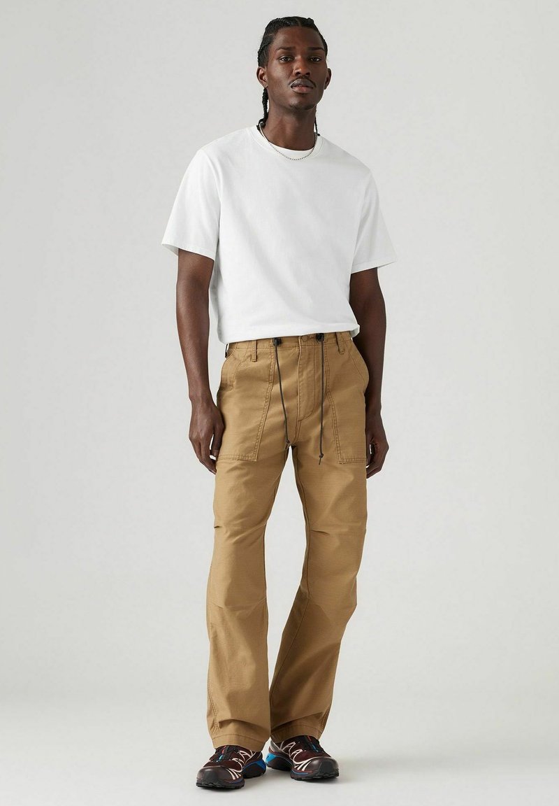 Camiseta blanca de algodón, corte holgado. Pantalones cargo marrones con cintura elástica y cordón ajustable, y bolsillos laterales. Zapatillas negras y rojas con detalles texturizados.