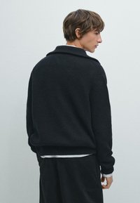 Zwarte gebreide trui met een brede kraag, mouwboorden en een relaxte pasvorm. Draag hem over een wit gestreept shirt, wat een gelaagde look laat zien.