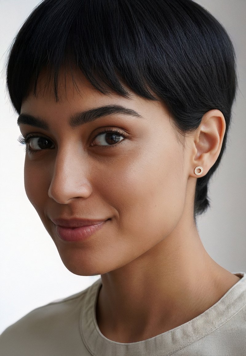 Rund guldstiftörring med cirkulär design och slät yta. Ämnet har kort mörkt hår och bär en ljusblå topp.