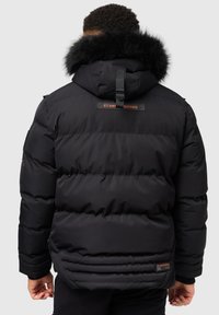Svart puffjacka med texturerad yta, som har en huva med fuskpäls, horisontell sömnad och en logotext nära nederkanten.