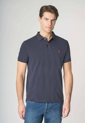 Giovane uomo che indossa una polo blu navy scuro con un logo ricamato rosso sul petto a sinistra e jeans blu, in piedi davanti a uno sfondo semplice.