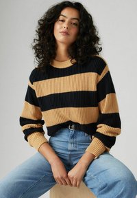 Maglione lavorato a maglia con strisce orizzontali nere e beige. Tessuto a coste, scollatura rotonda e vestibilità ampia. Indossato con jeans in denim a vita alta.