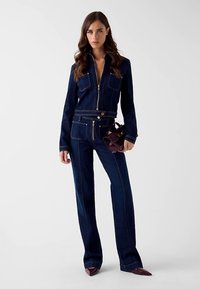 Dunkelblauer Jeans-Overall mit goldfarbenen Details. Verfügt über einen Reißverschluss vorne, zwei Brusttaschen und ausgestellte Hosenbeine. Akzente durch kontrastierende Nähte.