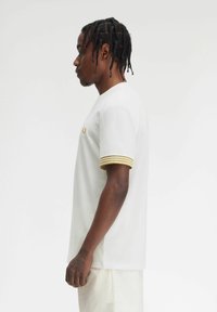 T-shirt bianca a maniche corte con tessuto testurizzato, dotata di polsini a righe gialle e un piccolo emblema sul petto. Design semplice, vestibilità regolare.