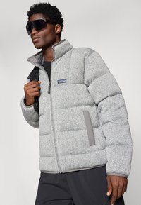 Šedá péřová bunda s vysokým límcem, s předním zipem, texturovanou látkou a kontrastními šedými akcenty na bocích.