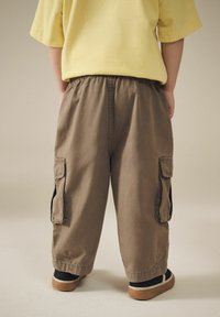 Brune cargo bukser med sidelommer, elastisk talje og en løs pasform. Bæres med sorte sko med en tan sål og en gul skjorte.
