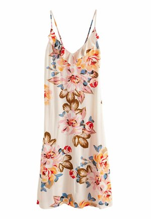 Femme en robe midi blanche à motifs floraux avec un col à volants et des sandales compensées noires, debout à côté d'un mur jaune vif et d'escaliers.