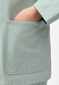 Jasnozielony materiałowy strój z dużą kieszenią. Materiał ma fakturowany wzór, a rękaw posiada prosty krój.