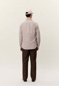 Mann iført beige stripete langermet skjorte, mørkebrune bukser, svarte sko og beige baseballcaps, vendt bort fra kameraet mot en nøytral bakgrunn.