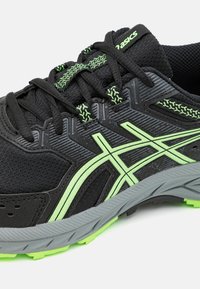 Zapatilla deportiva negra con suela gris, acentos verdes y parte superior texturizada. Presenta cordones y bordes reforzados para mayor durabilidad y soporte.