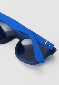 Kék napszemüveg matt felülettel, enyhén nagyobb, kerek lencsékkel és Ray-Ban logóval a szárakon. Textúrált, rugalmas karok.