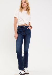 T-shirt blanc avec nœud à l'avant associé à un jean flair bleu foncé. Tenue complétée par des chaussures blanches avec des accents noirs et gris.