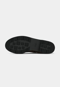 Chaussure en daim brun avec une semelle en caoutchouc noire. La semelle présente un motif texturé pour une meilleure adhérence, avec des éléments rectangulaires et circulaires.