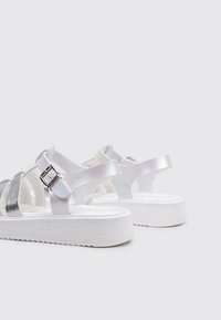 Sandalias blancas con tiras metálicas plateadas, suela plana de plataforma, hebillas ajustables y una superficie texturizada para un mejor agarre.