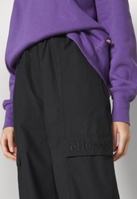 Sweat-shirt violet avec poignets côtelés, associé à un pantalon cargo noir ample avec un logo embossé sur la poche. Tissu en mélange de coton.