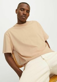 Lyse beige kortærmet T-shirt lavet af blødt bomuld, med rund halsudskæring. Båret med lyse bukser, siddende på en rattan stol.