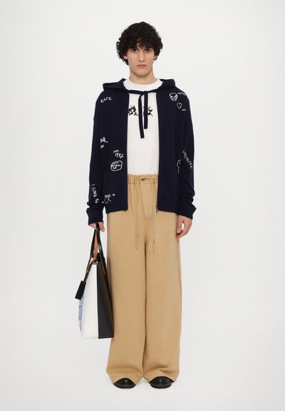 Sweat à capuche marine avec des motifs graphiques, chemise blanche avec détail de cravate, pantalon large beige et chaussures noires ; tenant un sac fourre-tout bicolore.
