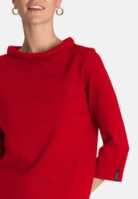 Blusa rossa realizzata in tessuto liscio, con scollatura a cappuccio e maniche a tre quarti. Dettaglio notevole include un piccolo logo sul polsino.