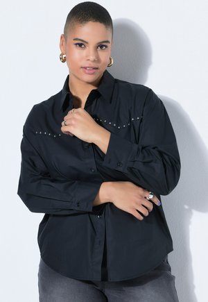 Person med kort hår iført en sort nittebeklædt skjorte og gyldne creoler, poserer med korslagte arme mod en hvid væg.