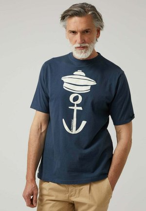 Hombre con cabello y barba canosos, vistiendo una camiseta azul marino con un gráfico de ancla blanca y un sombrero, combinada con pantalones beiges.