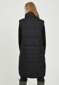 Gilet imbottito nero con colletto alto, texture trapuntata e design senza maniche, che arriva a metà coscia. Indossato su una maglietta a maniche lunghe.