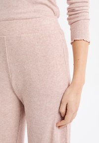 Pantaloni da soggiorno rosa chiaro a coste con una fascia larga in vita e una texture morbida. Vista ravvicinata che mostra una mano appoggiata sul tessuto.