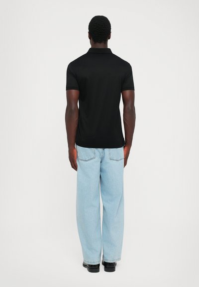 Bărbat stând cu spatele, purtând un tricou polo negru cu mânecă scurtă, blugi de culoare bleu deschis și pantofi negri, pe un fundal uniform.