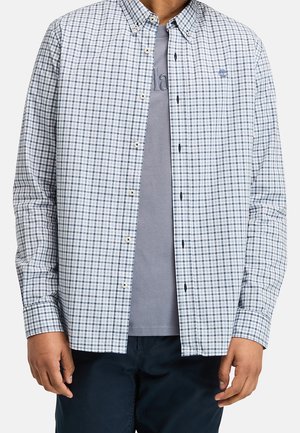 Mężczyzna w białej i niebieskiej koszuli w kratę noszonej na szarej koszulce oraz ciemnych spodniach, stojący z rękami luźno opuszczonymi wzdłuż ciała.