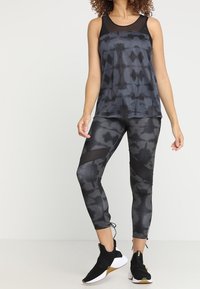 Kvinna klädd i en ärmlös topp med svart och grått batikmönster och matchande leggings med svarta meshpaneler, i kombination med svarta och vita träningsskor.