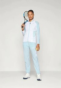 Frau in hellblauer und weißer Sportkleidung hält einen Tennisschläger über der Schulter und einen Tennisball in der linken Hand, während sie vor einem einfarbigen Hintergrund steht.