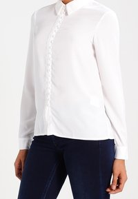 Donna che indossa una camicia bianca aderente con bottoni e maniche lunghe, insieme a jeans blu scuro, con le mani rilassate ai fianchi.
