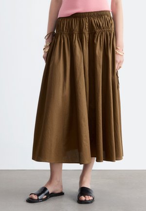 Femme portant une jupe midi marron à taille froncée, un haut rose, des sandales plates noires à bout ouvert et des bracelets dorés aux deux poignets.