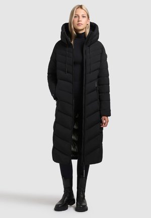 Donna che indossa un lungo cappotto imbottito nero con cappuccio, leggings neri e stivali neri, in piedi contro uno sfondo grigio chiaro uniforme.