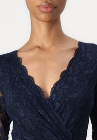 Vestido de encaje azul marino con un profundo escote en V, intrincado patrón floral y delicado borde festoneado en el escote y las mangas.